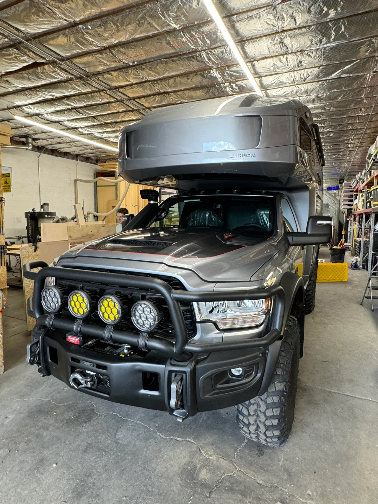 Front view of a gray composite camper on a regular-cab pickup inside a shop with foil-insulated ceiling and wood shelving.