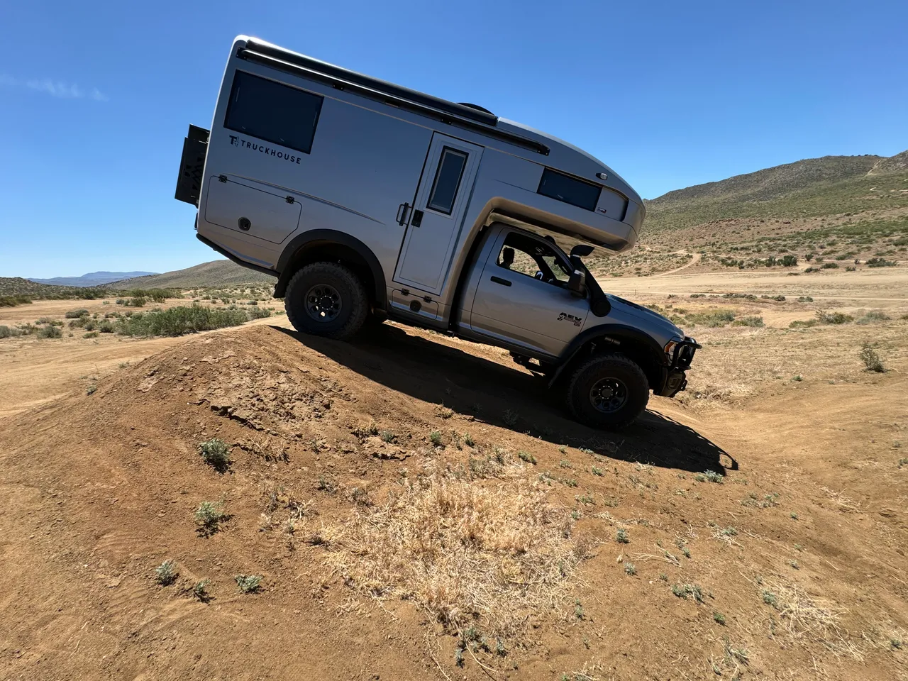 Side view of a silver camper on a regular-cab pickup climbing over a dirt mound in open desert, articulating the suspension.