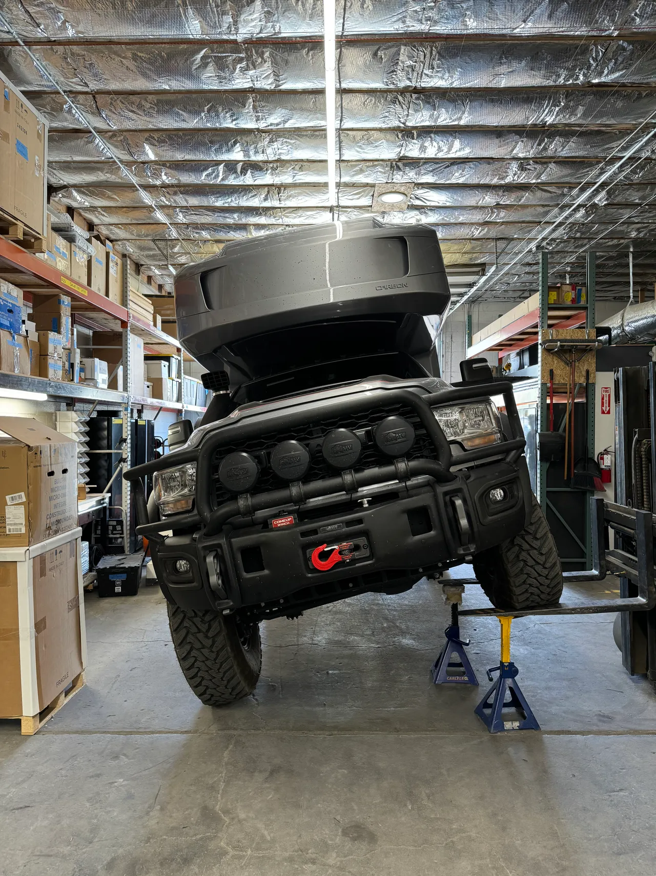 Gray camper truck in a shop with one front wheel lifted on a forklift and the frame supported on jack stands, twisting the chassis.