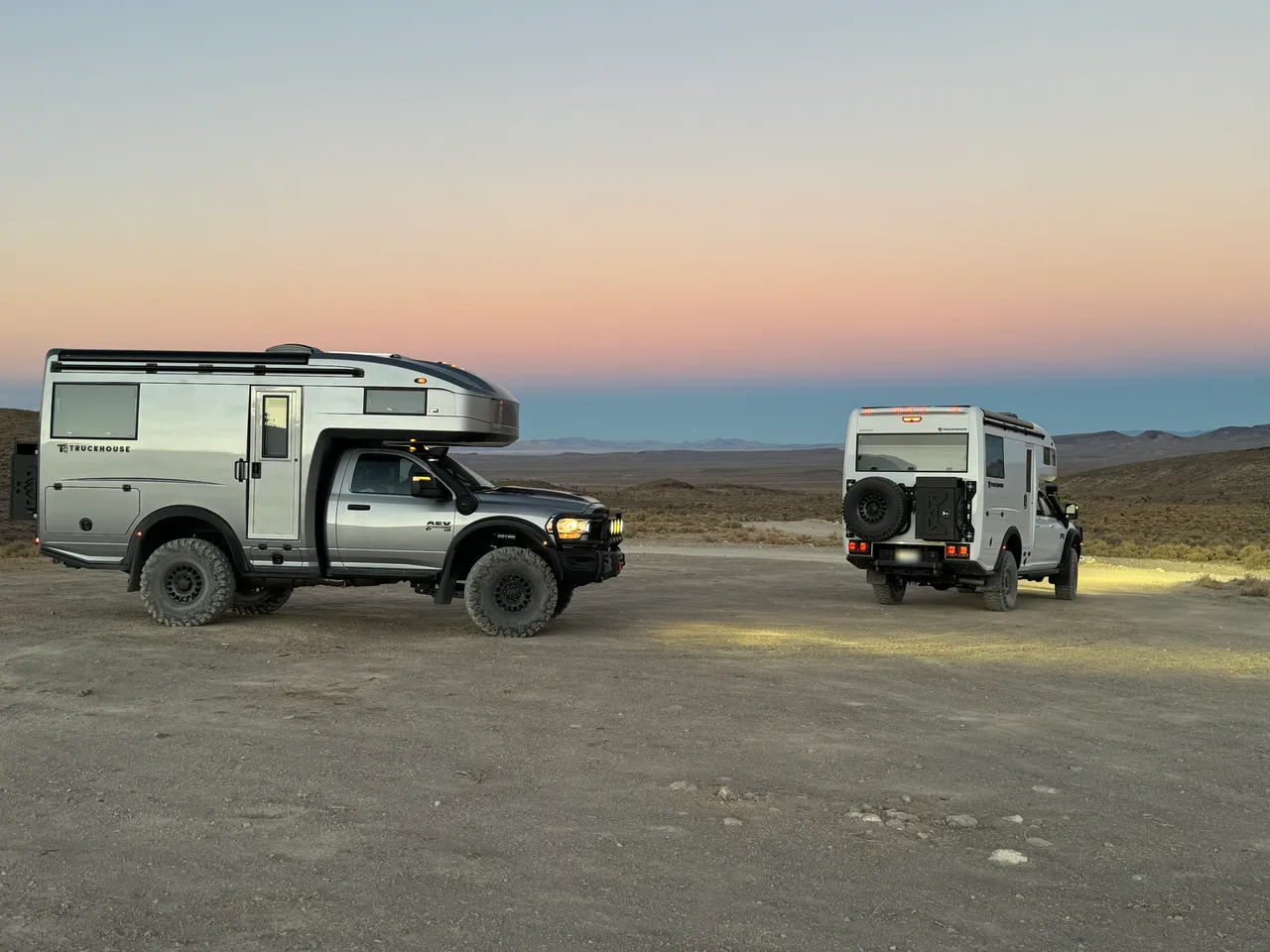 Two campers parked on a gravel desert flat at dusk: a silver one in side view and a white one from the rear with a swing-out spare tire carrier.