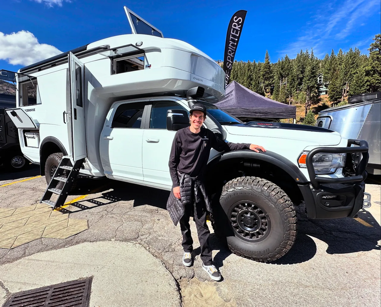 Petr standing beside a white composite camper mounted on a crew-cab pickup at an outdoor show, the camper door open and a fold-out entry stair deployed.