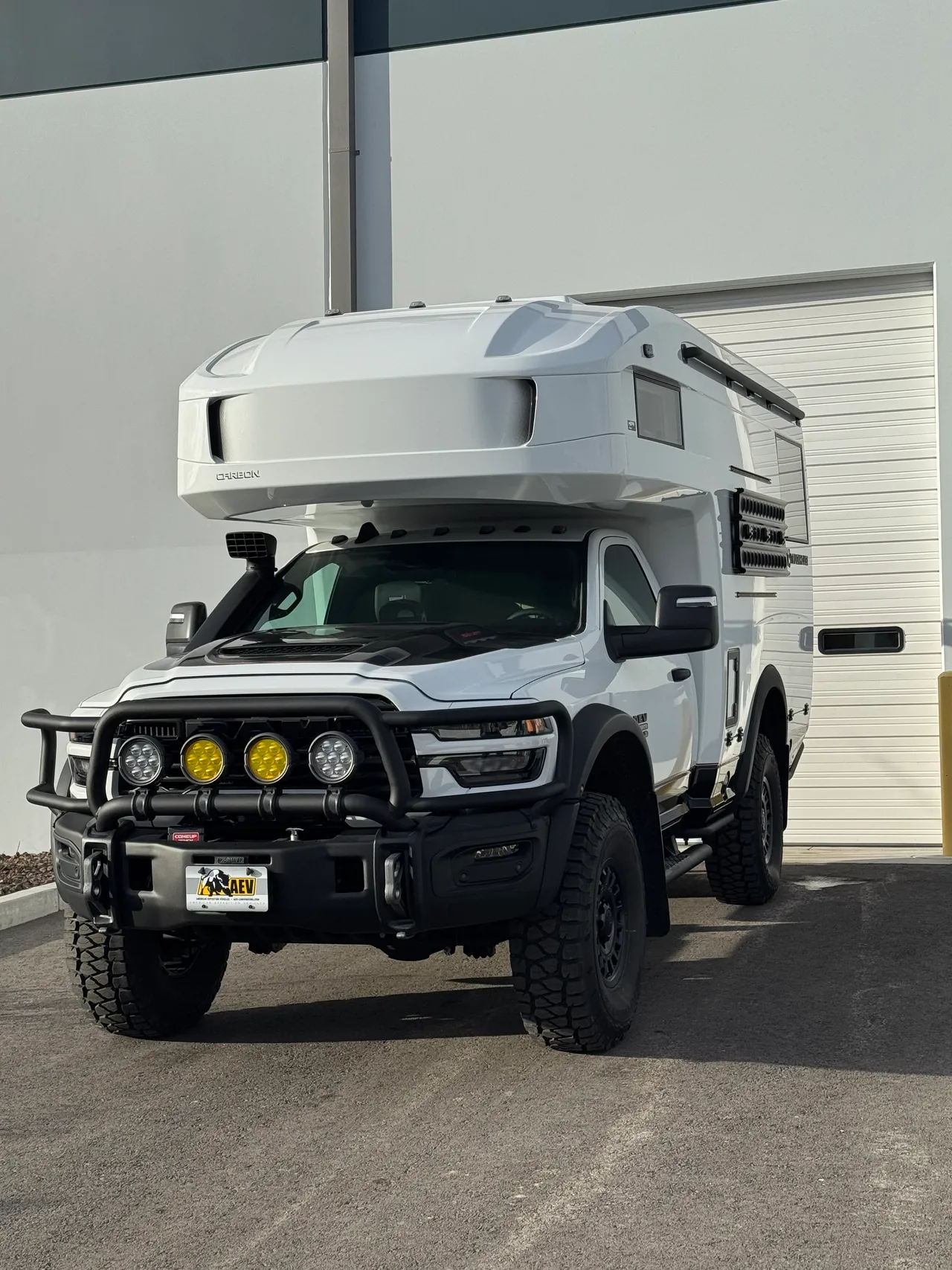 Front three-quarter view of a white composite camper on a regular-cab pickup parked in front of a white building, with four round auxiliary lights on the grille guard.