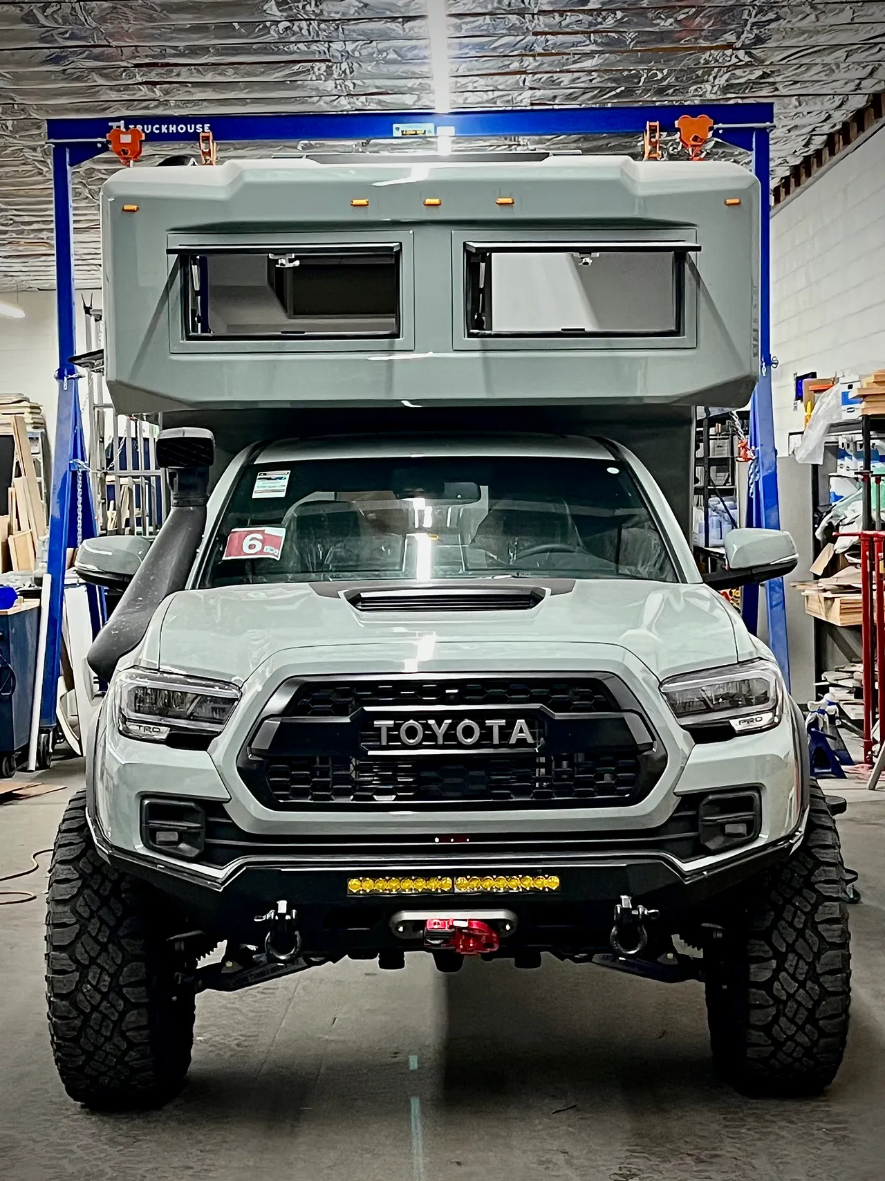 Head-on view of a gray camper on a mid-size pickup inside the shop, with two cab-over windows above the windshield.