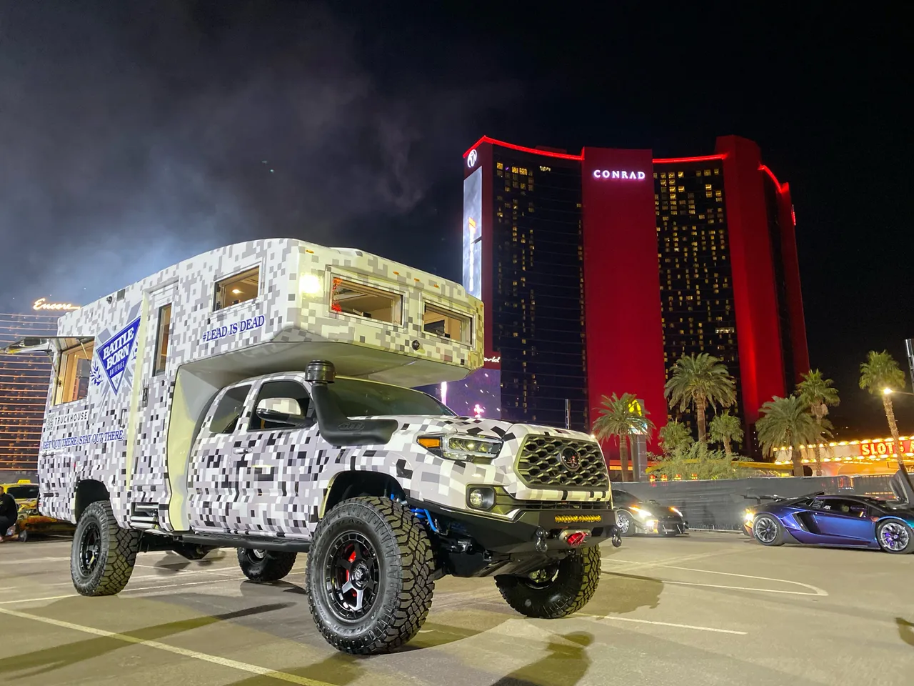 A camper on a mid-size pickup, both wrapped in a white and gray pixel camouflage pattern, parked at night in front of a red-lit hotel tower.
