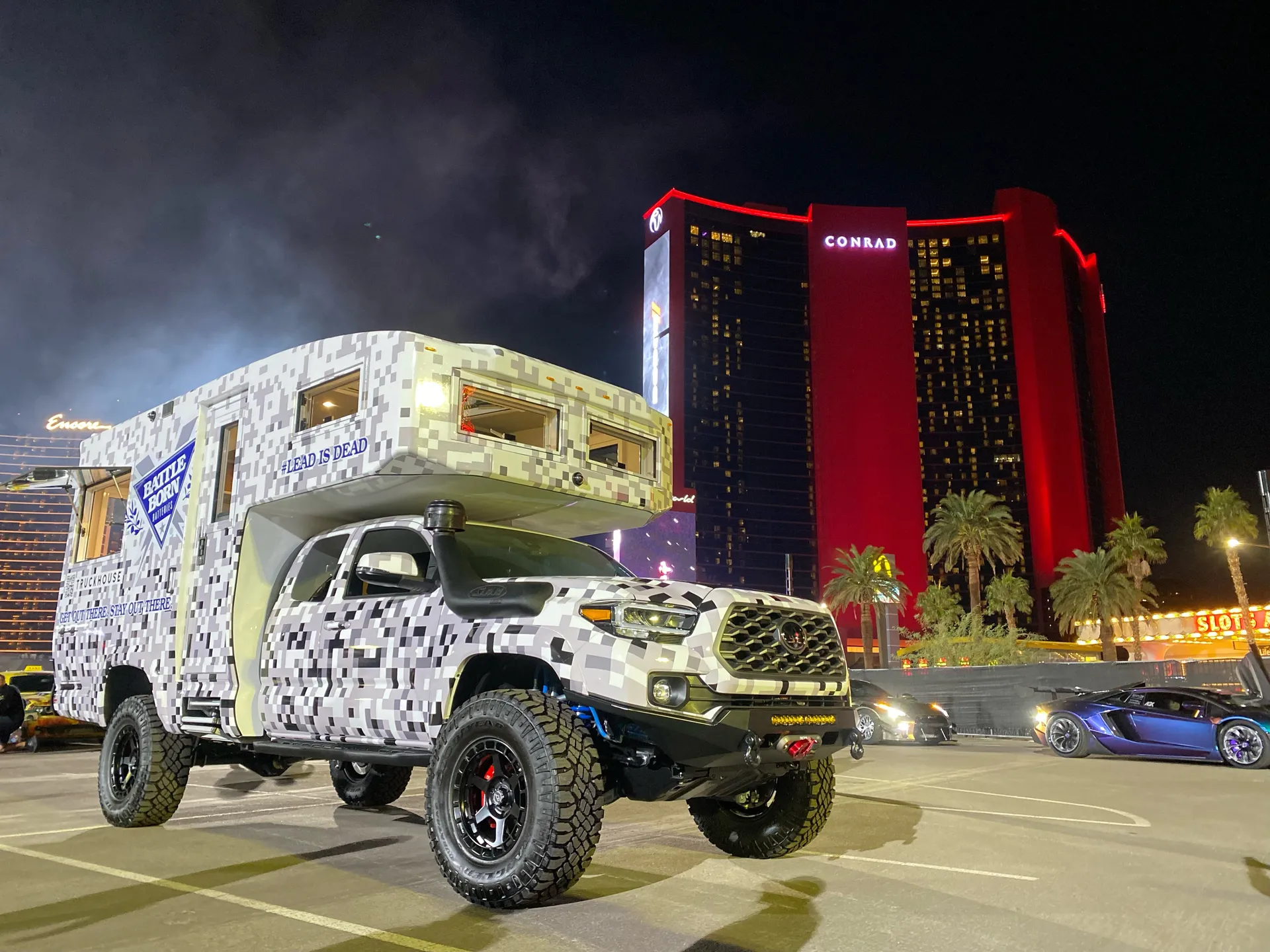 A camper on a mid-size pickup, both wrapped in a white and gray pixel camouflage pattern, parked at night in front of a red-lit hotel tower.