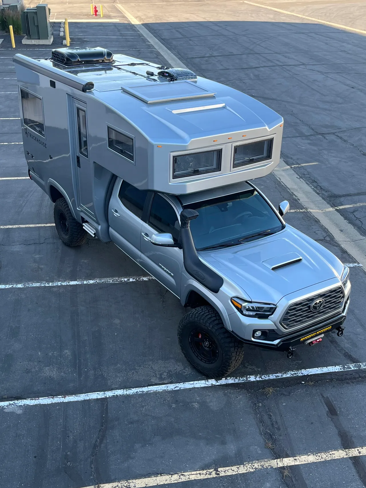 High overhead view of a silver composite camper on a mid-size crew-cab pickup in a parking lot, showing the roof hatch, vents and cab-over windows.
