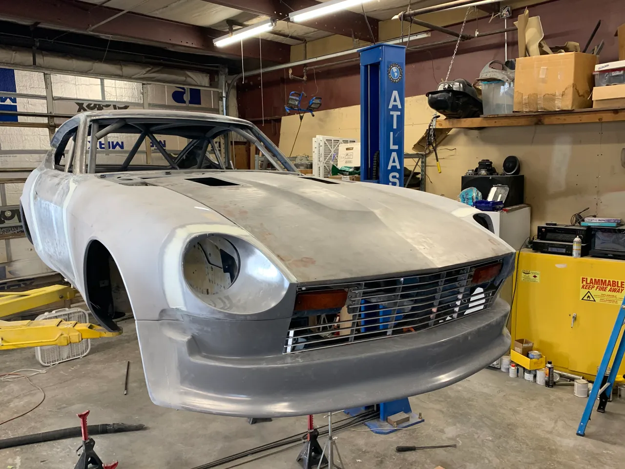 Front of the car in bare metal and gray primer with the new front air dam and grille fitted, parked in front of a blue two-post lift.