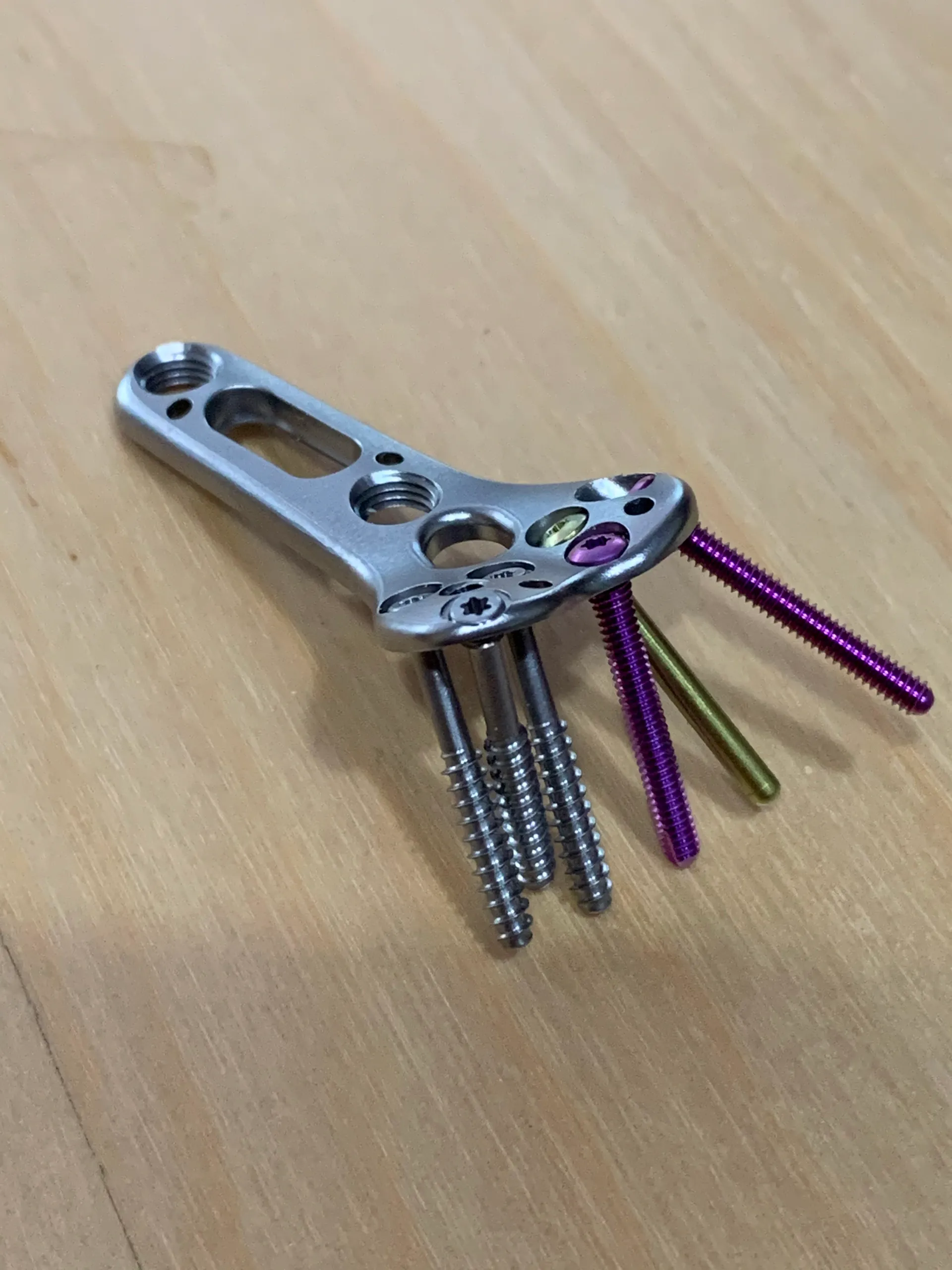 A first-article distal radius plate on a wooden surface with silver locking screws and purple and gold threaded pins passing through its head.