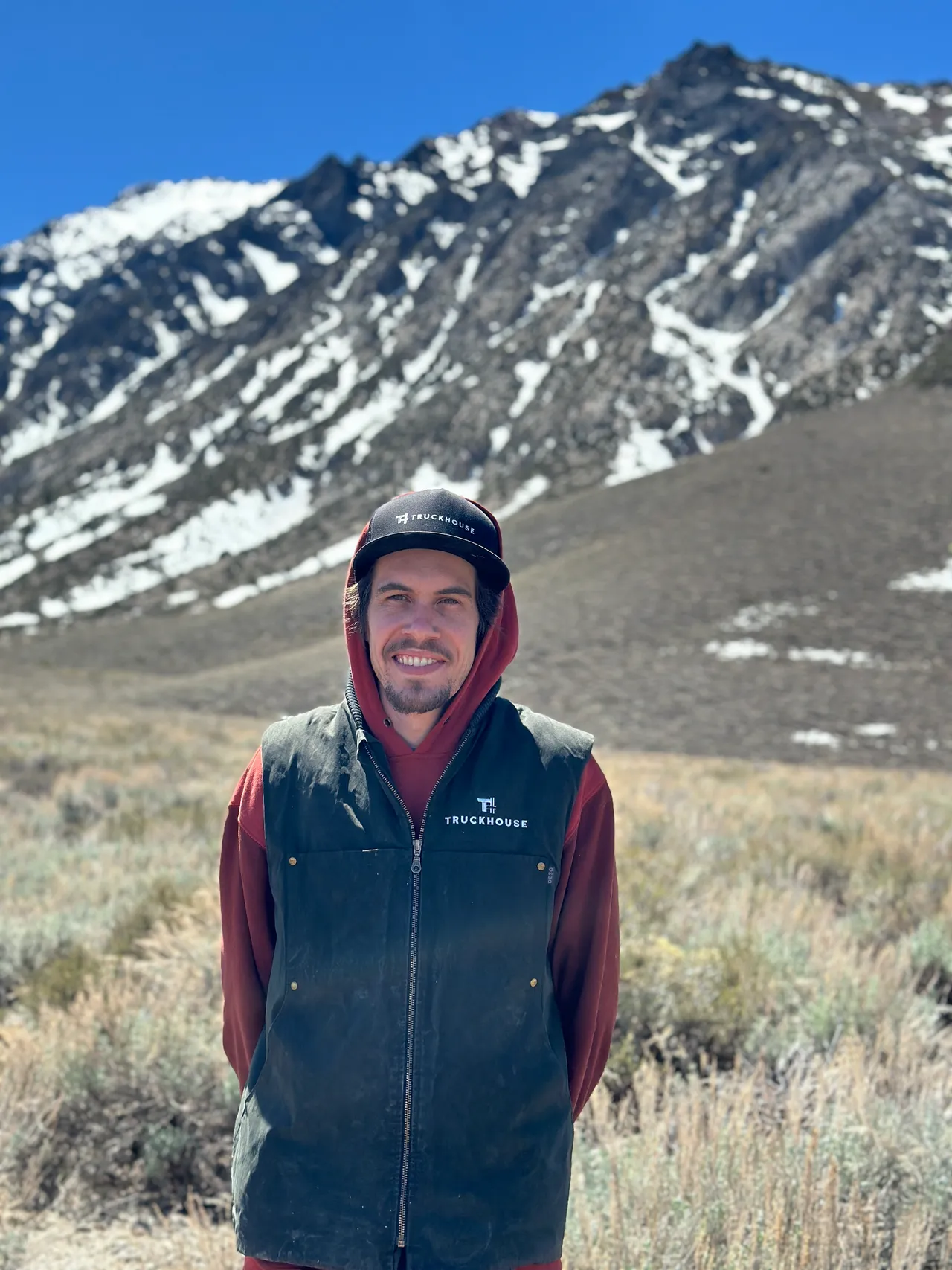 Petr smiling in a Truckhouse cap and vest, standing in sagebrush below a snow-streaked mountain