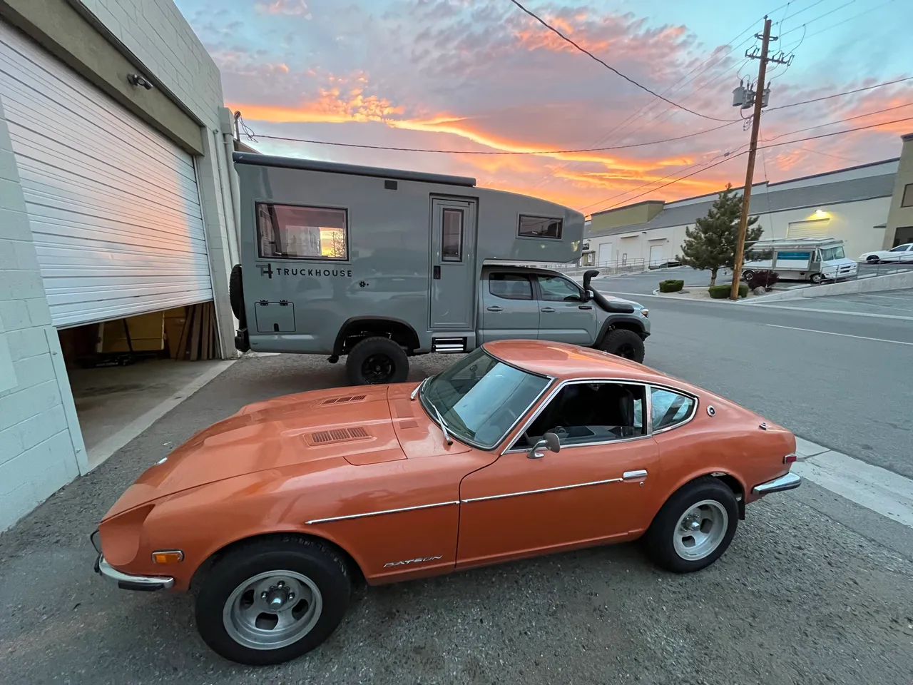 The orange 240Z parked outside a shop at sunset in front of a gray camper-equipped pickup.
